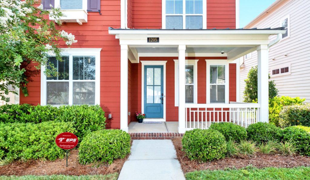 Close up of a new two-story red home with white trim, a blue front door, and a small yard with bushes and a small front porch. The EPS Security sign is out front.