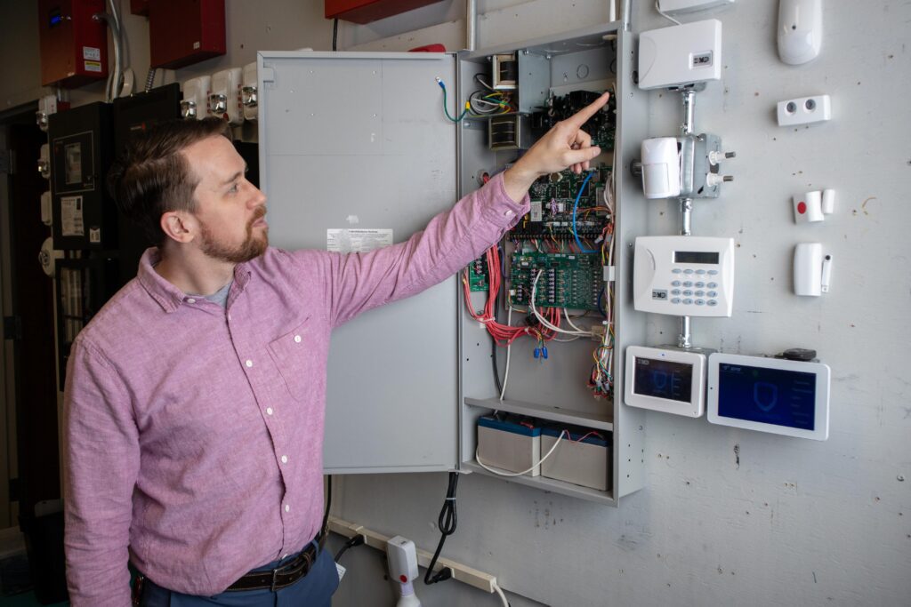 Tony standing and pointing/teaching about a security panel with the panel open showing the electical parts within the box.